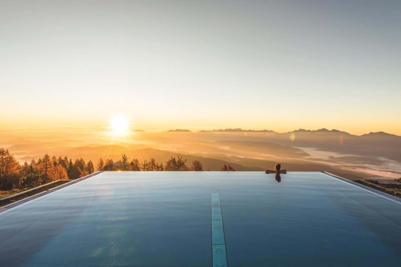 Unendlichpool bei Sonnenaufgang, eine Person an der Poolkante, darunter Nebelmeer und Bergketten